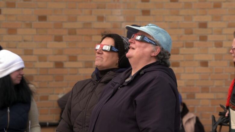 Two women are wearing solar eclipse glasses and look at the sky.