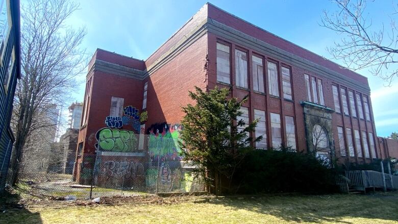 A red brick two-storey building has green shrubs in the front yard, with chain link fence. There is colourful graffiti on the left side of the facade