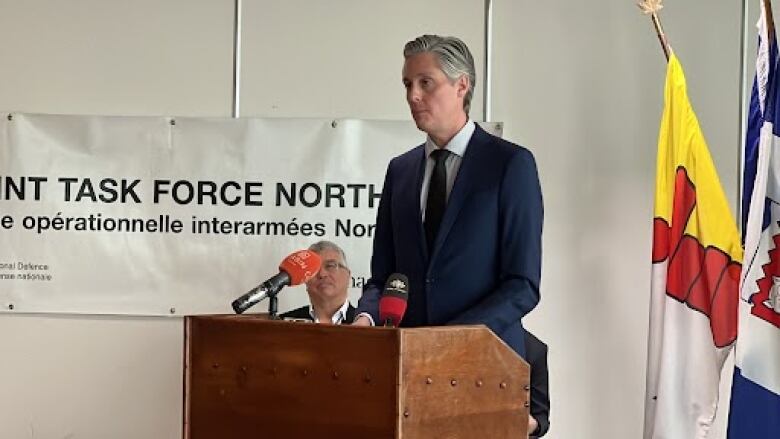 A man stands at a podium with flags and a sign in the background. 