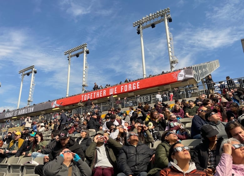 People sit in the stands of a football stadium looking up at the sky with eclipse glasses on.