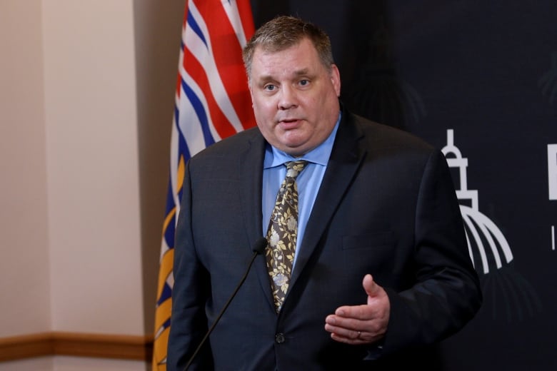 A white man wearing a blue shirt and a patterned black tie speaks in front of a B.C. flag.