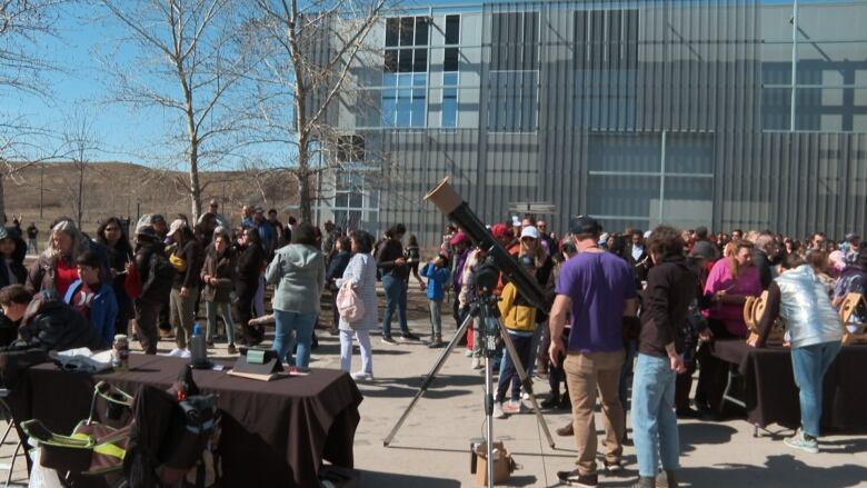 A crowd is gathered on a sidewalk, around telescopes.