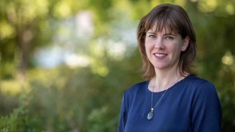 A woman in a blue sweater standing in front of some blurry greenery. She is smiling at the camera, has shoulder-length light brown hair and is wearing a necklace