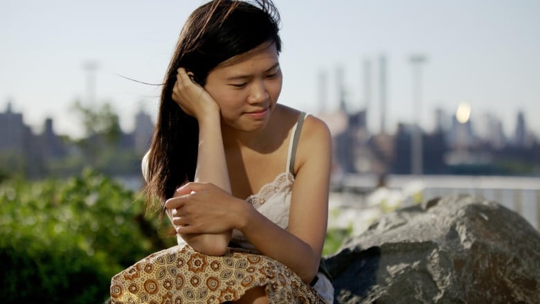 An Asian woman with long black hair with her head resting on her hand. The skyline of Toronto is blurry in the background.