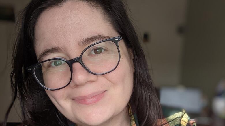 A close-up portrait of a woman with dark hair and black-rimmed glasses who is smiling softly at the camera.