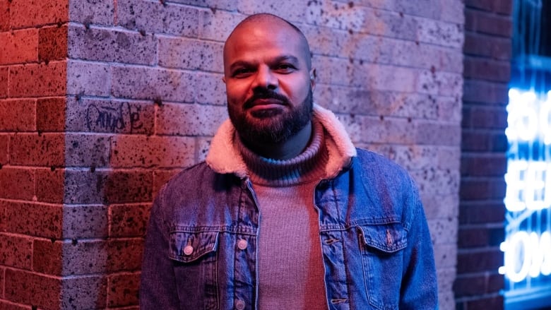 A bearded man wearing a jean sherpa jacket over a taupe turtleneck and standing in front of a brick wall.