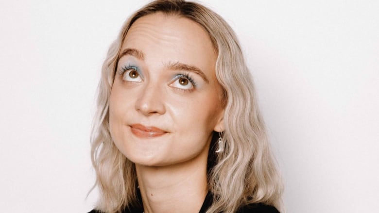 A blonde woman with shoulder-length hair who is looking up while standing in front of a white background.