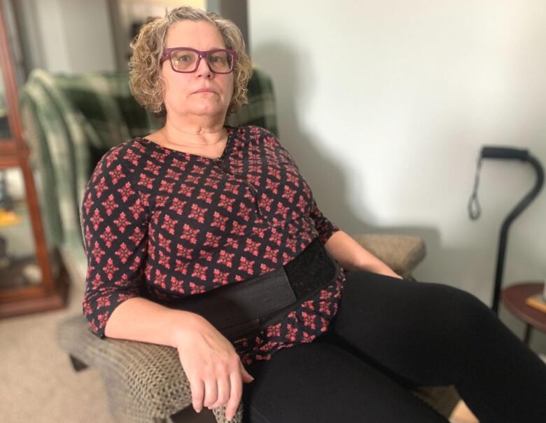 A woman in a patterned blouse and black pants sits in an easy chair. Behind her, a cane rests against the wall.