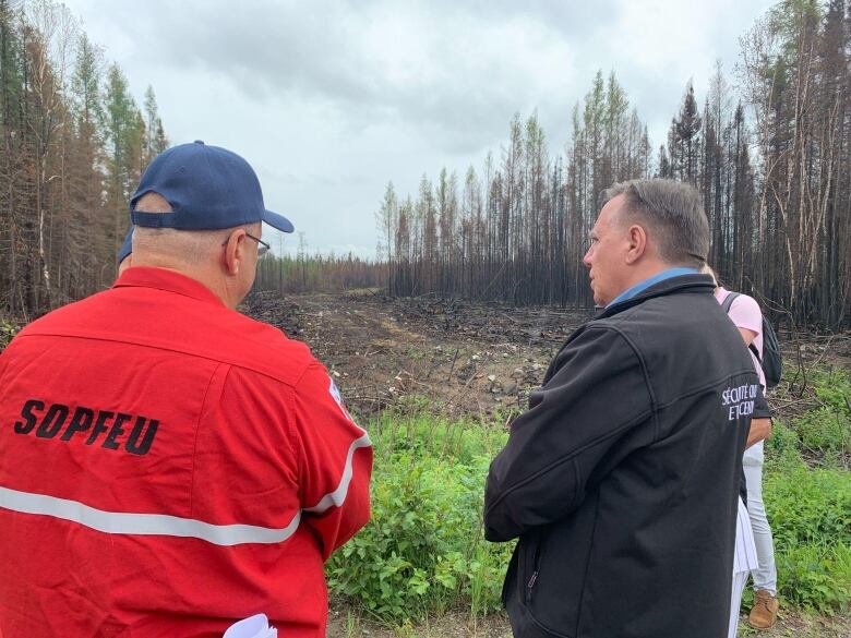 A man with a jacket that says SOPFEU stands talking to another man. They are looking on a field and a forest with burned trees.