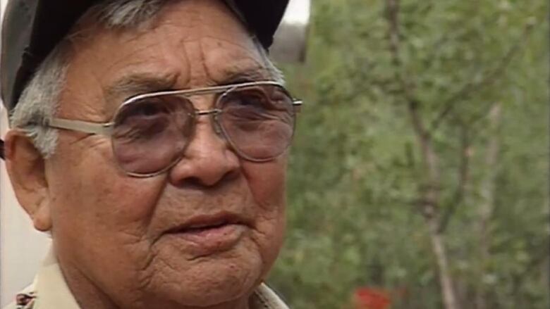 A older man in glasses and a hat speaks to the camera. 