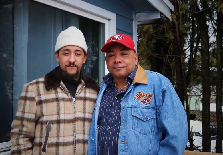 Two men stand side by side, staring at a camera on a deck.