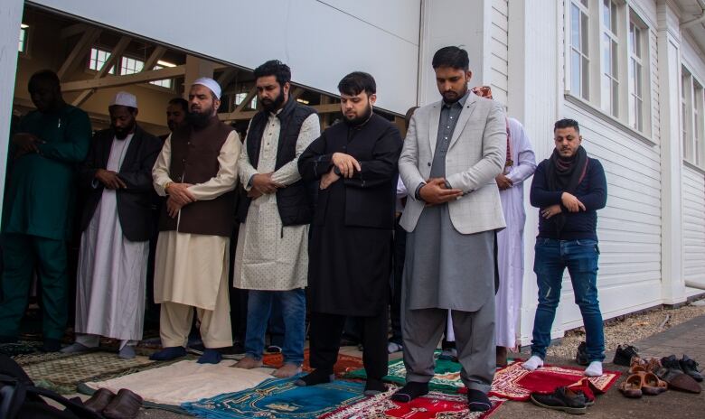 A group of men stand, heads bowed in prayer, outside of a building