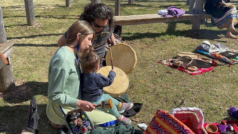 A woman and a man holding hand drums sit on grass with a baby, who's lifting a drum stick over the drum.