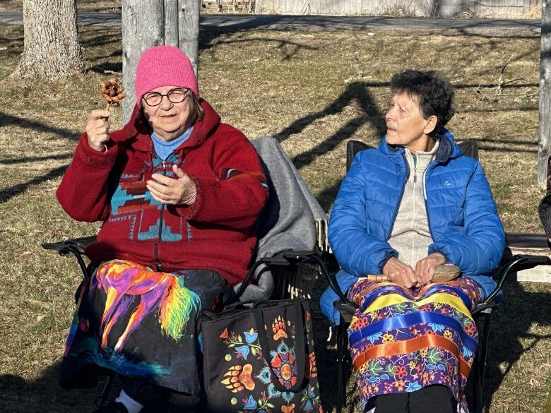 Two elderly women wearing long, colourful skirts and bundled up in winter coats and hats sit in lawn chairs in the sun.