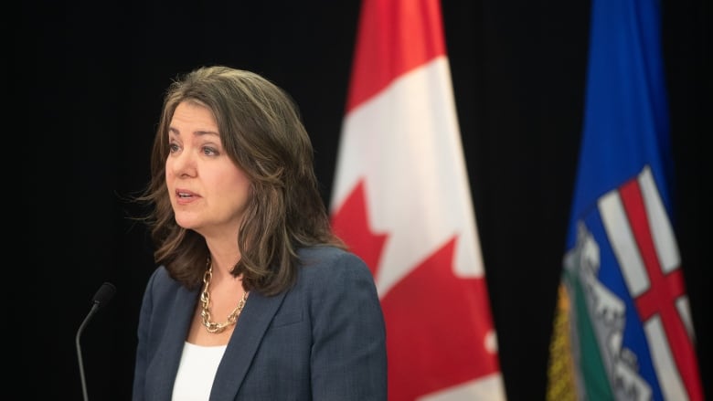 a woman speaks into a thin microphone, with Canada and Alberta flags behind her.