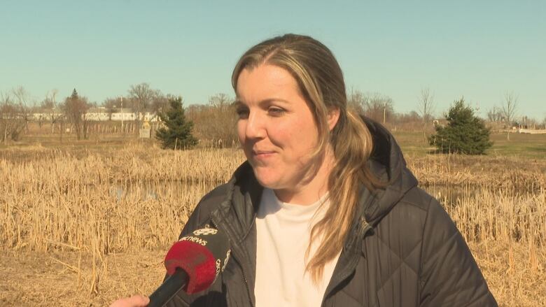 Keisha Rose Topic stand for an interview outside in a white shirt and black jacket. There is a small pond, grass and trees behind her. 