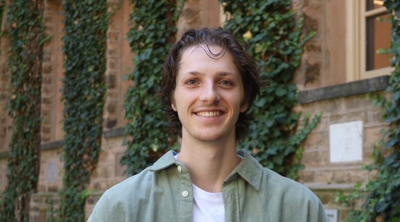 A portrait of a person in an open green button-up standing before a brick wall.