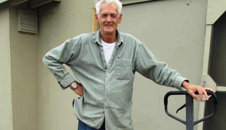 A man with white hair in a celadon shirt leans on the handle of a hand truck