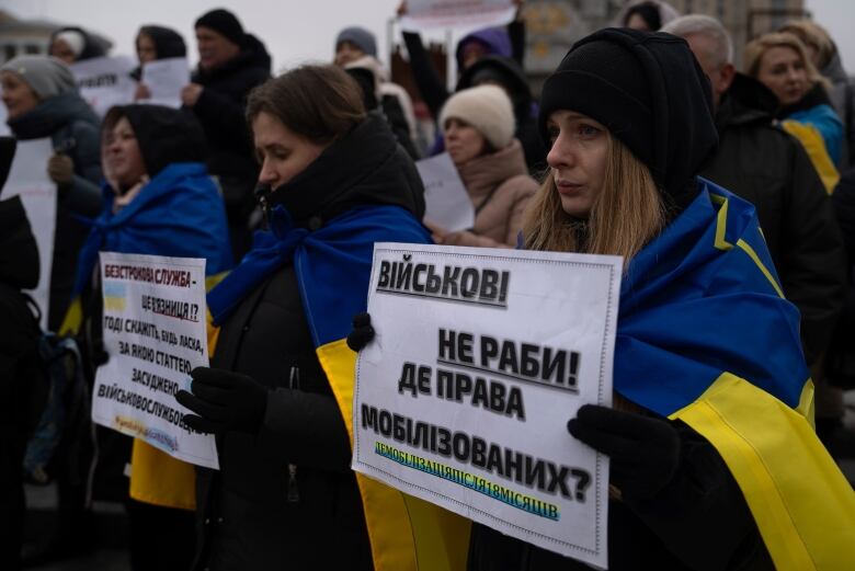 Women stand outside in winter clothes, with blue and white Ukrainian flags draped over their shoulders, holding signs with Ukrainian text on them. 