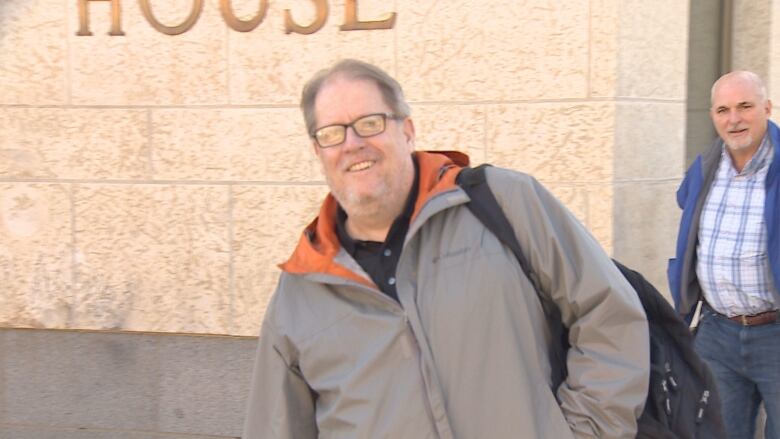 A man in a grey coat smiles in from of a courthouse. 