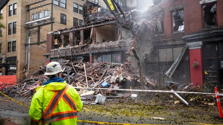 A building being demolished as a worker in reflective clothing looks on.