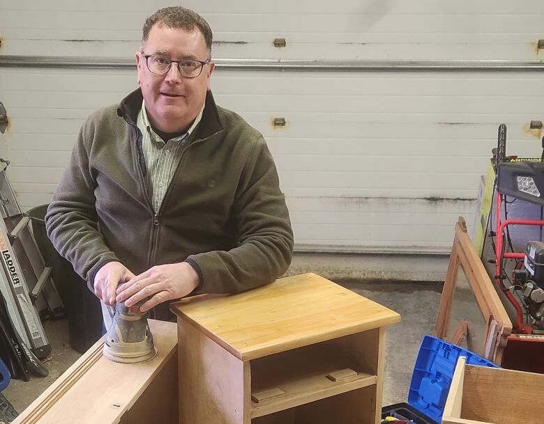 A man looks at the camera while sanding a piece of furniture.