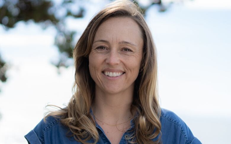 A woman with long light-brown hair and wearing a dark blue, short-sleeved denim top, smiles broadly for a head and shoulders portrait against a backdrop of out-of-focus clouds, sky and leaves on an overhanging tree branch.