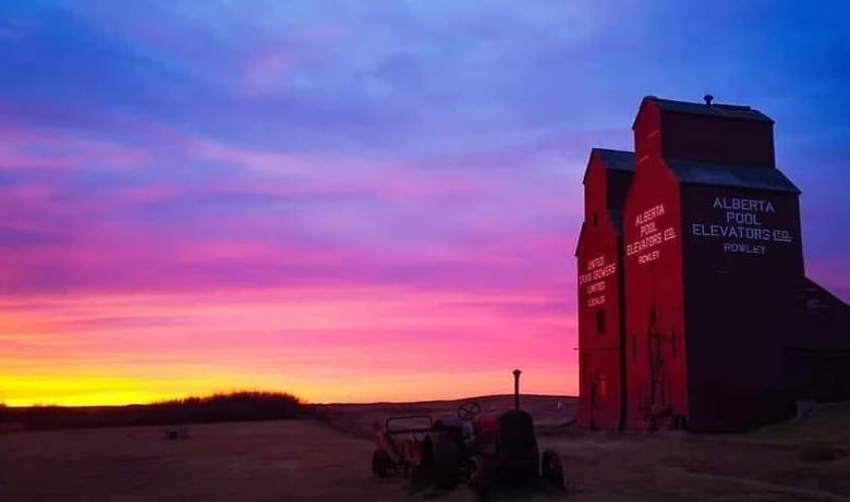 two wooden grain elevators stand in a field at sunset.