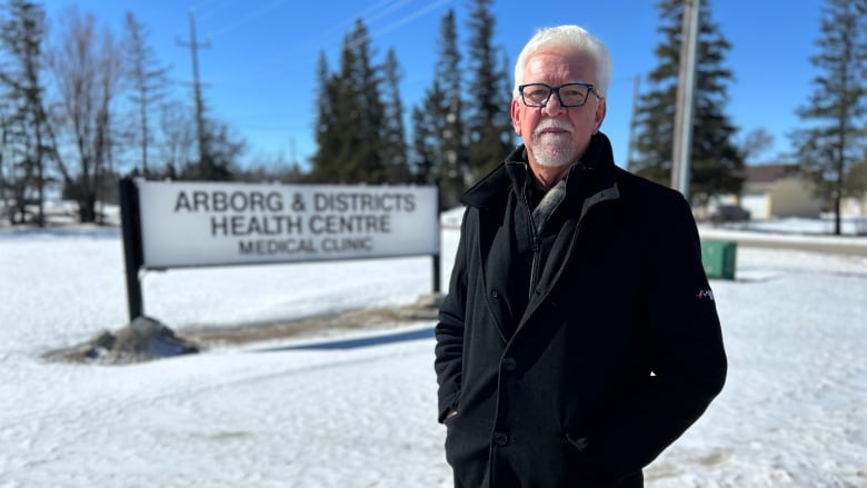 A man wearing a black jacket and pants stands in front of a sign that says Arborg & Districts Health Centre.