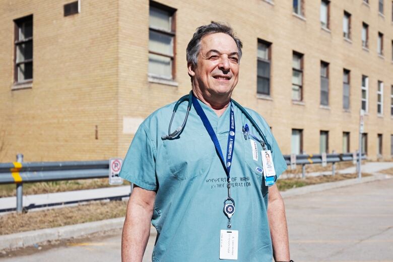 A doctor wearing blue scrubs with a stethoscope around his neck is pictured in front of a brick building with windows on a sunny day.