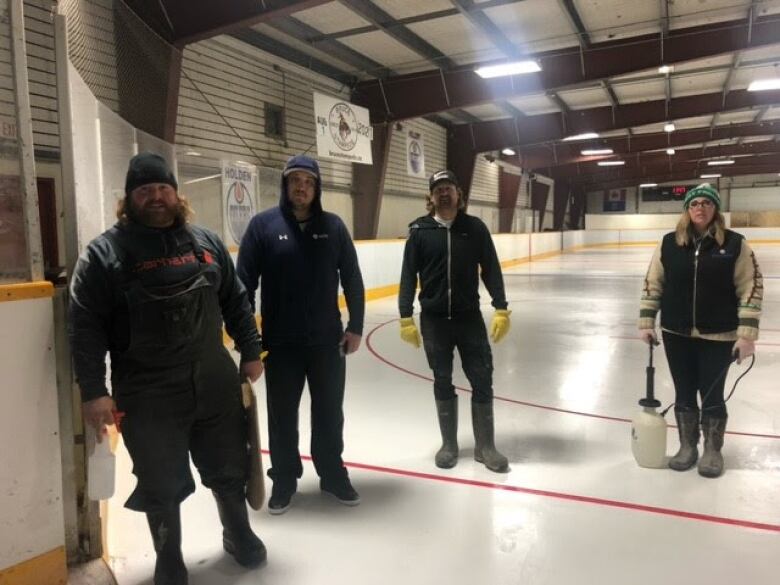 Three men and one woman are standing inside a community hockey arena. They are on the ice, near the boards, smiling toward the camera. They are in street clothes.