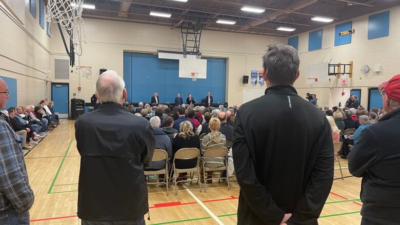 A crowd attends a public meeting in a school gymnasium.