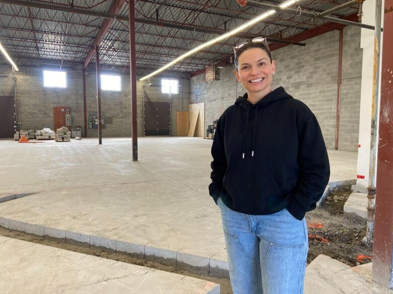 a white woman wearing jeans and a black sweater stands in a large warehouse, which will soon be a food bank with a kitchen.