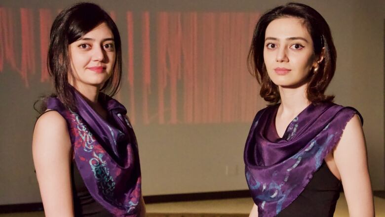 Two women with olive skin, long dark hair and dark blue shawls draped over their chests stand facing each other but looking at the viewer.