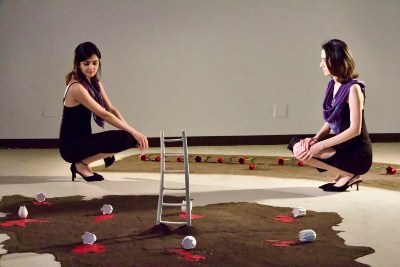 Two young women on opposite sides of the photo sit on their haunches, in high heels, amidst their artistic display of a map covered in red with a ladder sitting atop it. 