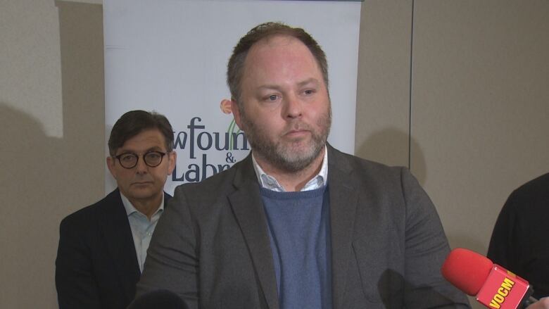 A man in a sweater and suit jacket stands in front of a Newfoundland and Labrador government banner.