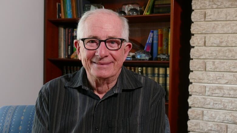 James Parker is a 78 year old caucasian man wearing a grey striped shirt and black framed glasses. He's sitting on a blue striped chair in front of a bookshelf. You can see part of a brick fireplace to his left. He has a slight smile on his face.