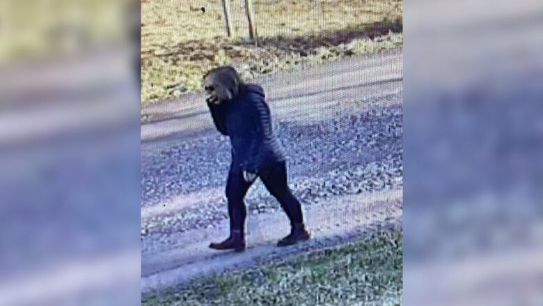 A woman wearing a dark puffer coast and black pants is seen walking along a gravel road.