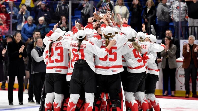 A team of women's hockey players celebrate in a group while raising a trophy.