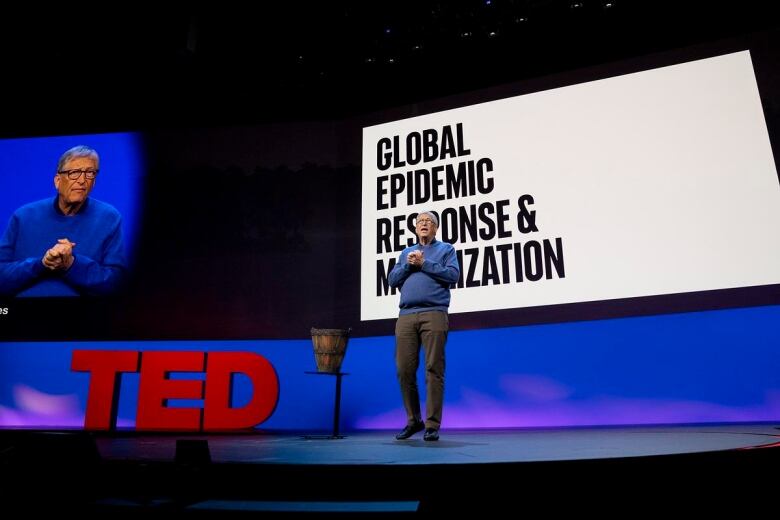 A man in a blue sweater with glasses speaks from a stage.