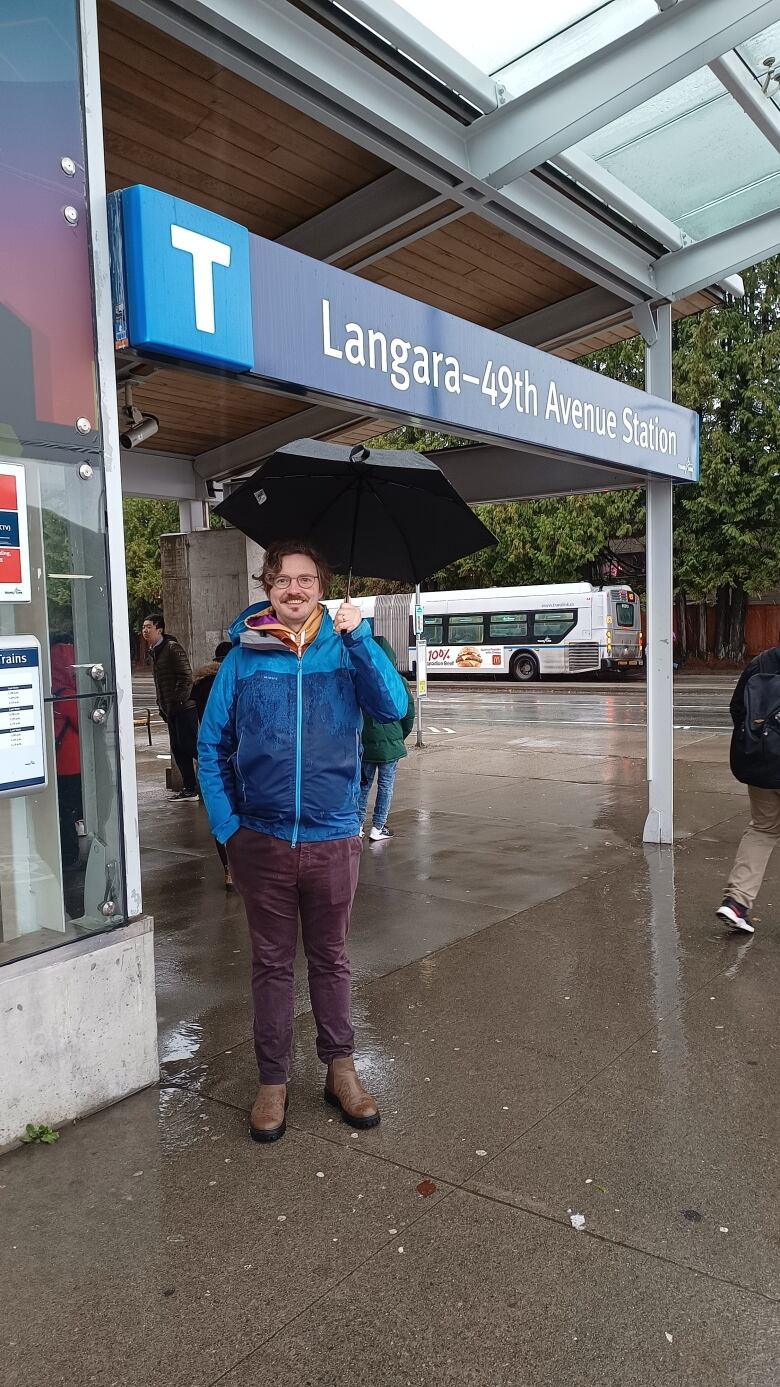 A white man carrying an umbrella is pictured standing outside a SkyTrain station.