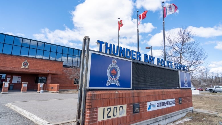 Flags are seen at the entrance of a red brick building. The sign says Thunder Bay Police Services. 