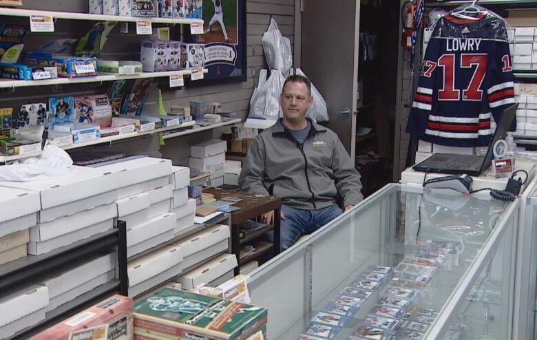 A man sits behind a display counter which features trading cards.