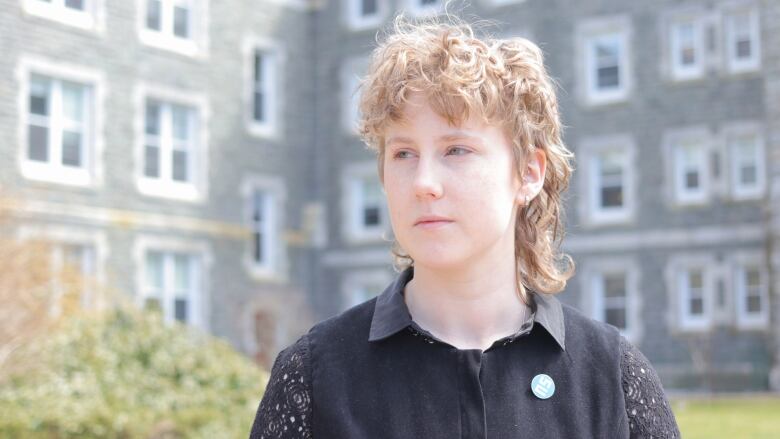 A non-binary person with curly hair and a mullet-type hairstyle is standing in front of a stone university building. They're wearing a collared shirt.