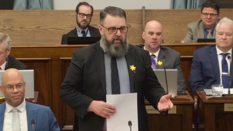 A man with a black suit and a beard stands up and questions the Health Minister.