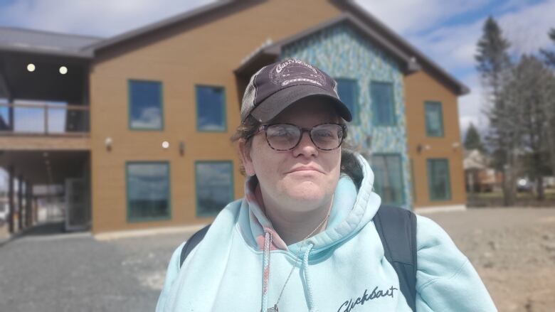 A woman wearing a baseball cap, glasses, a light blue sweatshirt and a backpack smiles while standing in front of a building under constructed. 