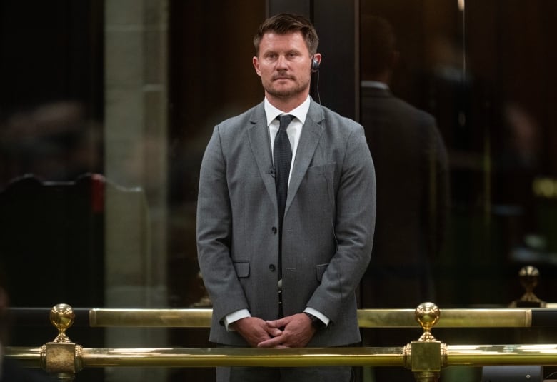 A man in a grey suit stands in front of the bar of the House of Commons.