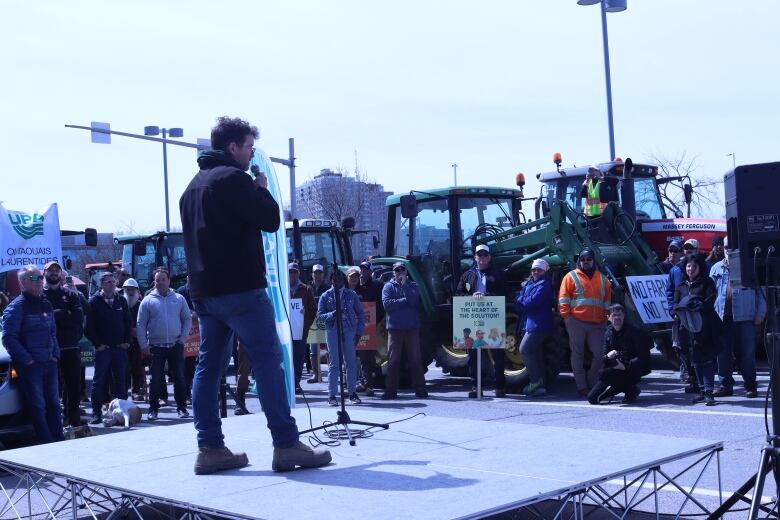 A speaker stands on a stage in front of a crowd of protestors and their tractors.