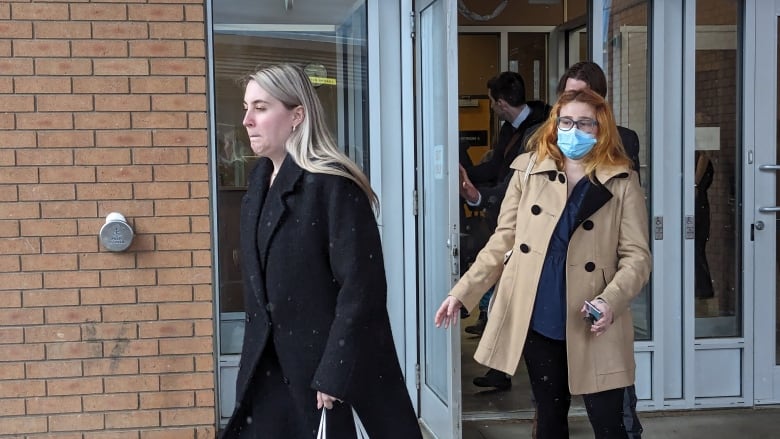 A woman in a beige jacket and orange hair walks out of a building behind a blond haired woman in a black jacket