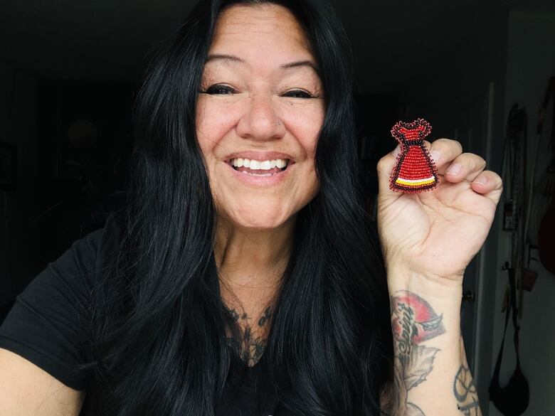 A smiling woman with long, dark hair holds up a miniature red dress made of beads.